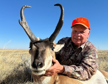 A proud hunter wearing an orange cap and camouflage shirt poses with a trophy antelope, displaying its prominent horns, during a successful hunt with SNS Outfitter & Guides.